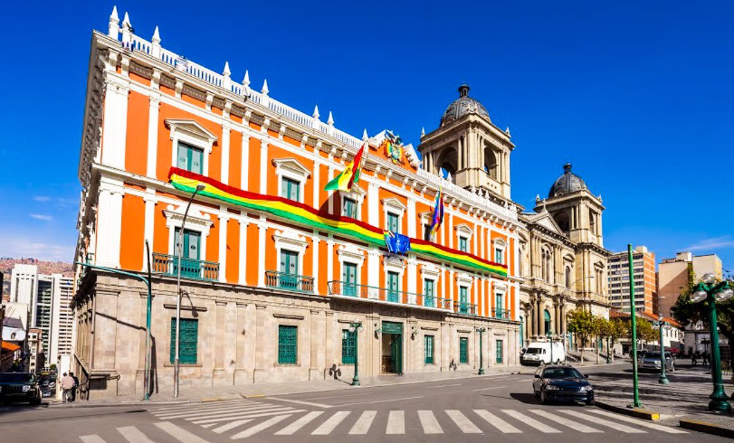 A large, orange and white neoclassical building with green-trimmed windows, adorned with Bolivian flags and a multicolored banner, stands at a street corner in La Paz under a clear blue sky.