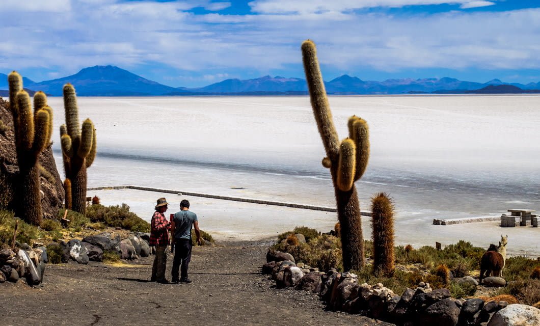Two people walk along a rocky path bordered by large cacti, overlooking the vast white expanse of Salar de Uyuni salt flats—one of the top Bolivia highlights—while distant mountains frame the blue sky.