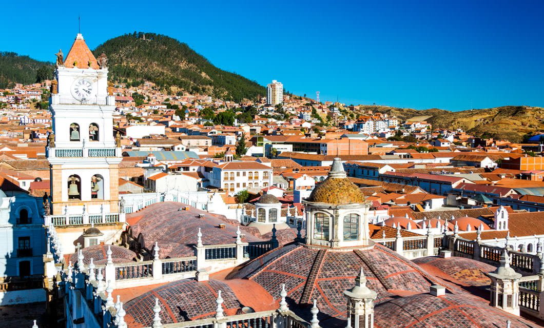 Rooftop view of a city with red-tiled roofs, a clock tower, mountainous landscape, and clear blue sky—capturing the charm of a historic Latin American town on a Classic Bolivia Tour.