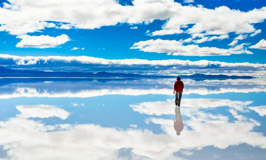 A person in a red jacket walks on the vast reflective surface of Uyuni, mirroring the blue sky and white clouds, with distant mountains on the horizon.