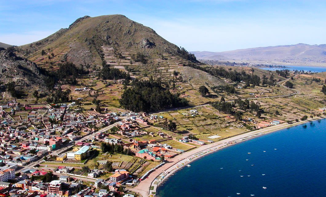 A coastal town with colorful buildings sits beside a curving bay with calm blue water and boats, making it a perfect stop on a Coast to Coast Tour, all backed by terraced hills and a tree-covered mountain under a clear sky.