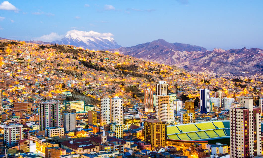Colorful cityscape of La Paz, Bolivia, as seen on a Classic Bolivia Tour, with densely packed buildings on hilly terrain, a stadium in the foreground, and snow-capped mountains rising under a clear dusk sky.
