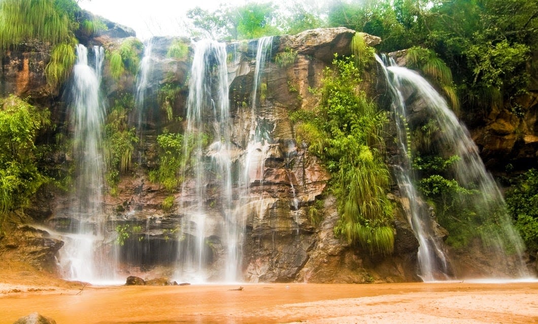 A wide waterfall cascades down a rocky cliff covered with green plants, surrounded by lush vegetation—one of the Bolivia highlights you’ll encounter on your Bolivia travel adventure. The water flows into a shallow, muddy pool at the base.