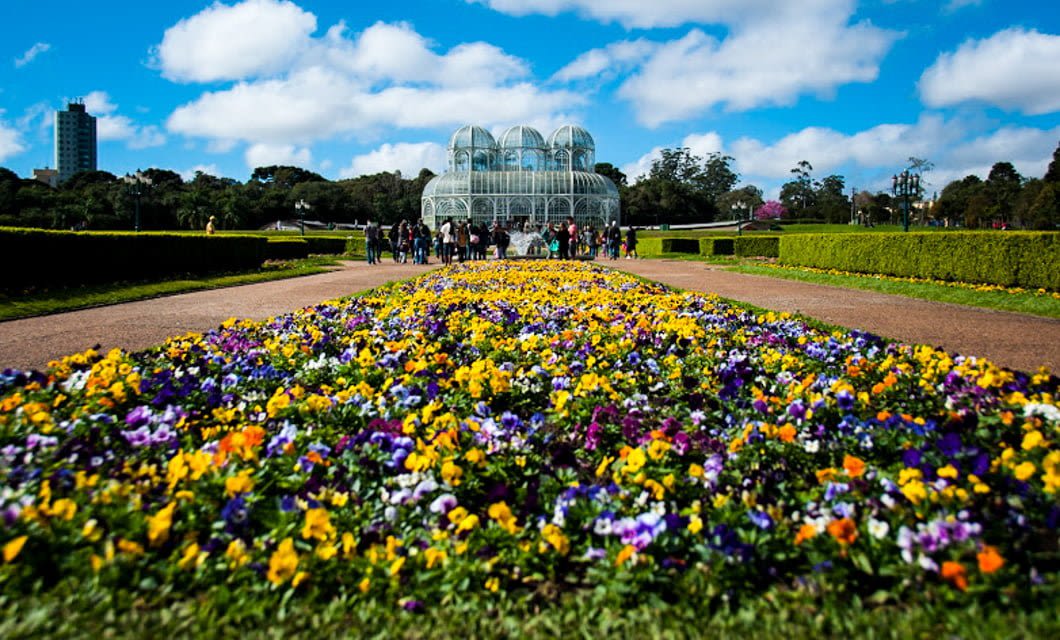A wide flowerbed with colorful flowers leads to a glass greenhouse in a botanical garden, surrounded by green lawns and hedges—perfect for relaxing after a scenic Brazil Train Tour under a blue sky with scattered clouds.