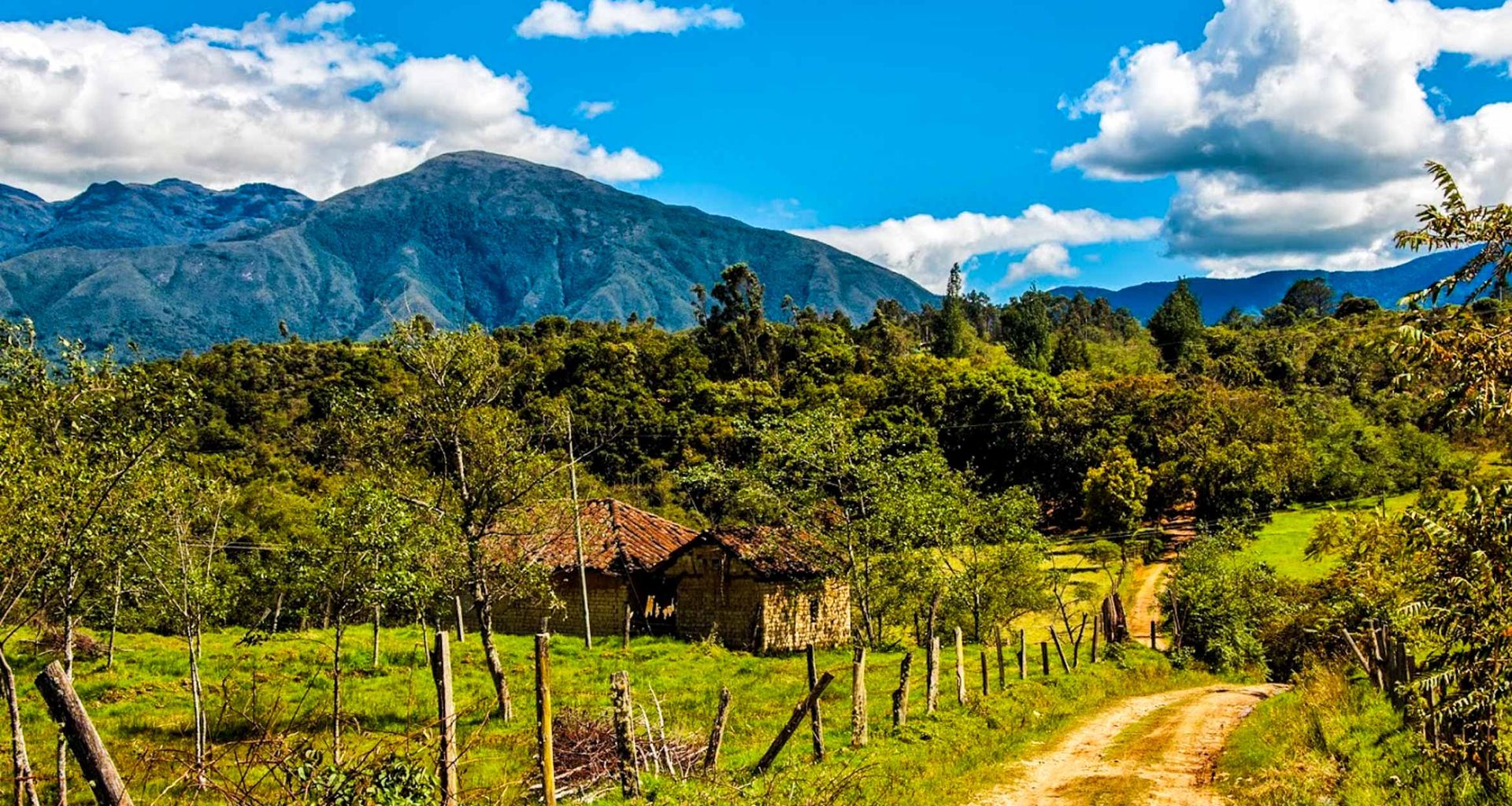 A dirt road winds through green fields and trees toward a rustic house with a red-tiled roof, set against forested hills and the tall, blue Boyaca mountain range of Colombia under a partly cloudy sky—a perfect scene for a scenic tour.