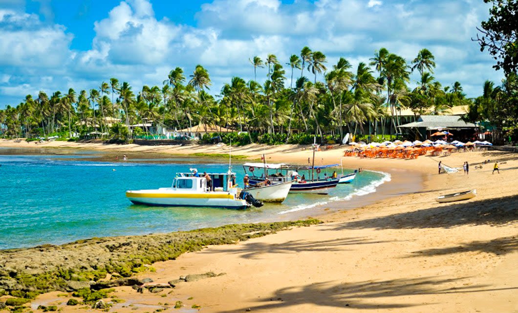 A tropical beach scene with boats anchored on the sand, clear blue water, and palm trees lining the shore—reminiscent of Chapada Diamantina’s beauty, where diamonds in nature await beyond umbrellas and lounge chairs under a partly cloudy sky.