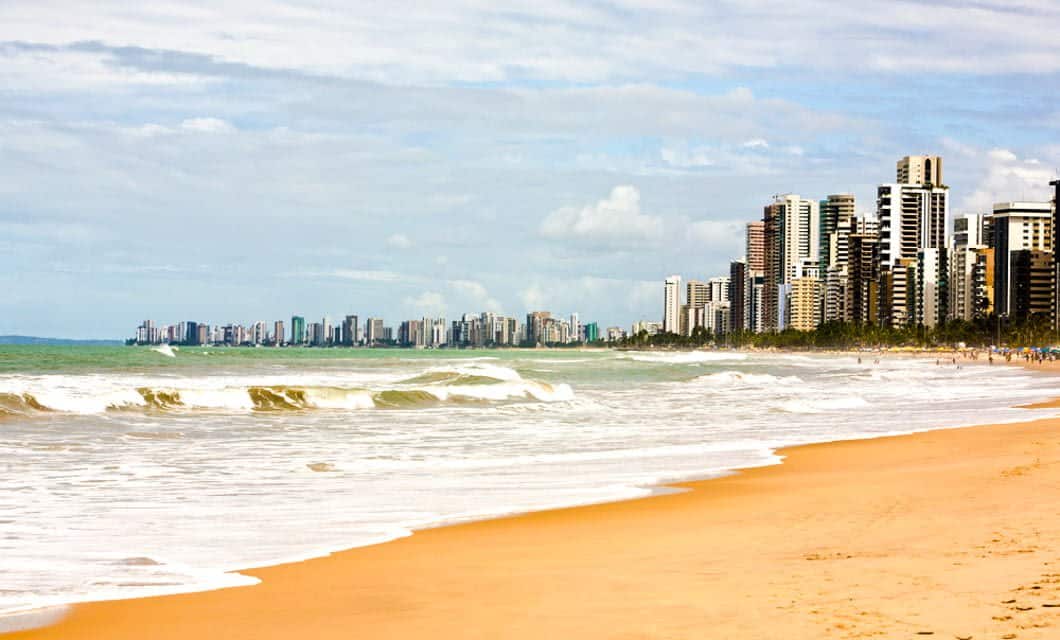 A sandy beach with gentle waves and turquoise water, the Coast of the Conquerors is lined with tall modern buildings and skyscrapers under a partly cloudy sky.
