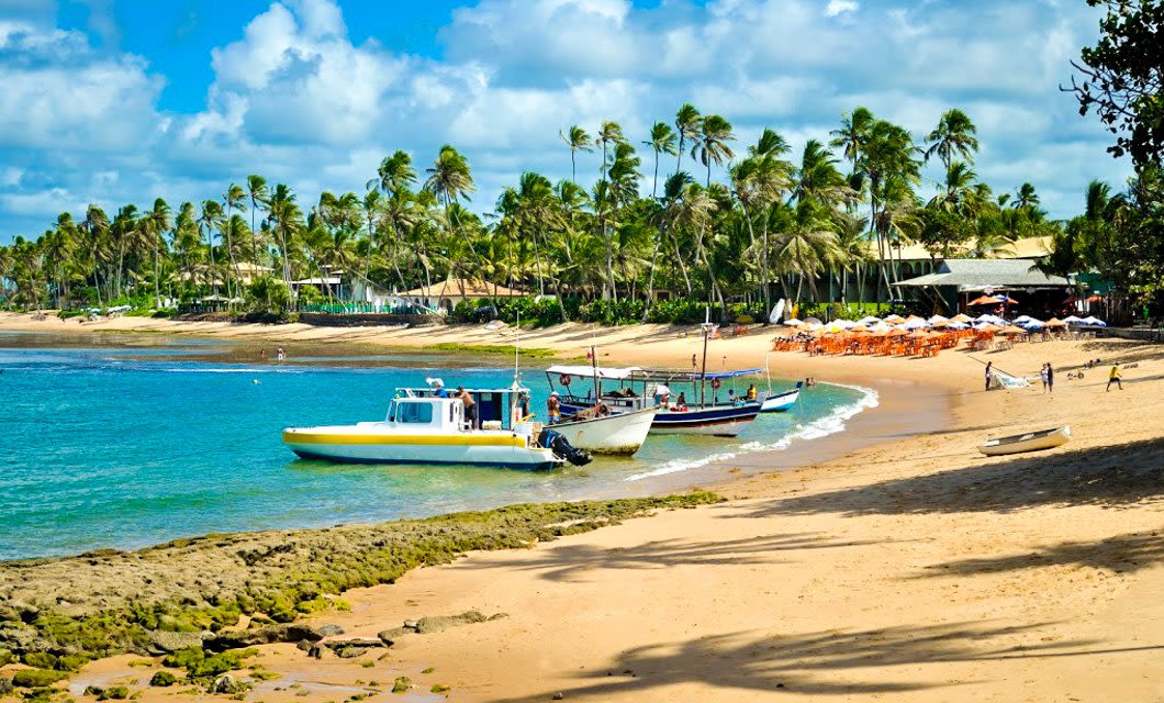 A sandy tropical beach with several boats anchored near the shore, palm trees lining the coastline, and beach chairs with umbrellas in the distance—perfect for relaxing after a Brazil Golf Tour under a bright blue sky with scattered clouds.