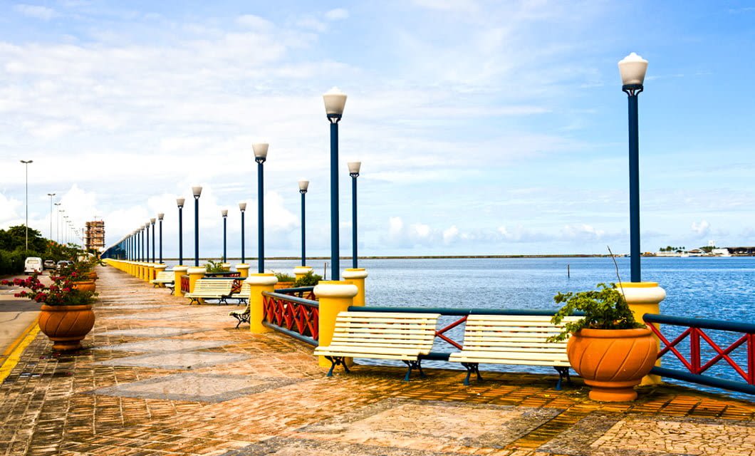 A scenic waterfront promenade along the Coast of the Conquerors, with rows of lampposts, yellow benches, and large potted plants, overlooks a calm blue body of water under a partly cloudy sky.