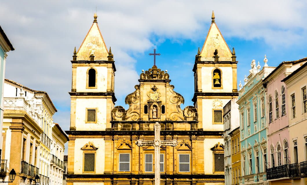 Historic yellow stone church with two towers and a central cross, flanked by colorful colonial buildings under a partly cloudy sky on the Coast of the Conquerors.