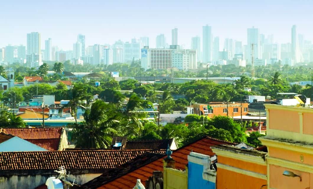 A colorful neighborhood with tiled roofs and lush trees sits in the foreground, set along the Coast of the Conquerors, with tall modern skyscrapers rising against a hazy sky in the distance.