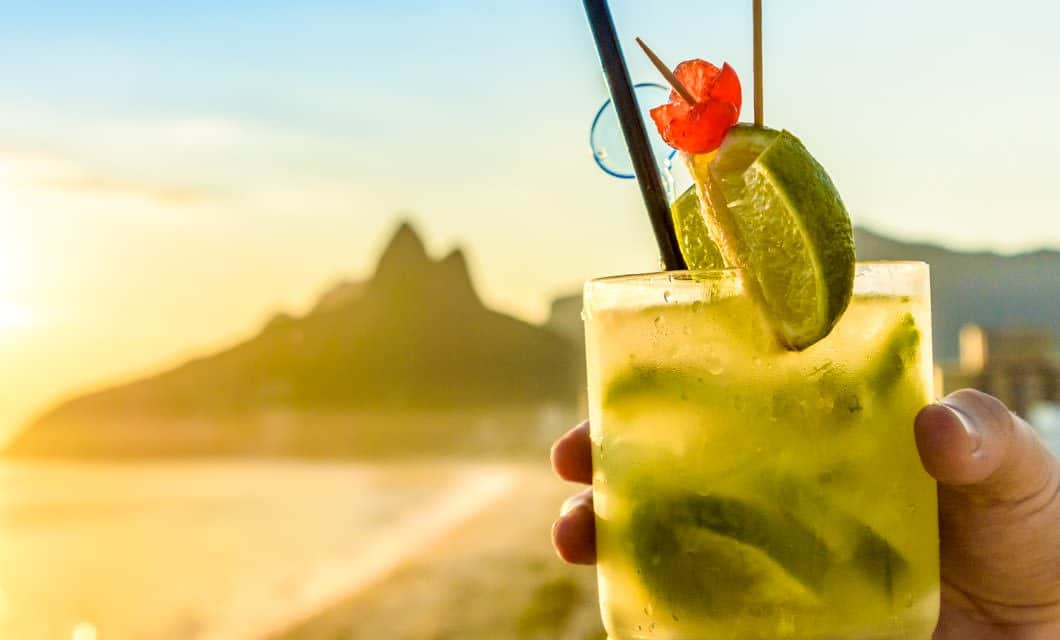 A hand holds a cocktail glass filled with lime slices, ice, and a cherry garnish, with a straw. This refreshing drink is set against a sunny beach and mountain backdrop at sunset, capturing the vibrant spirit of Brazilian cuisine.