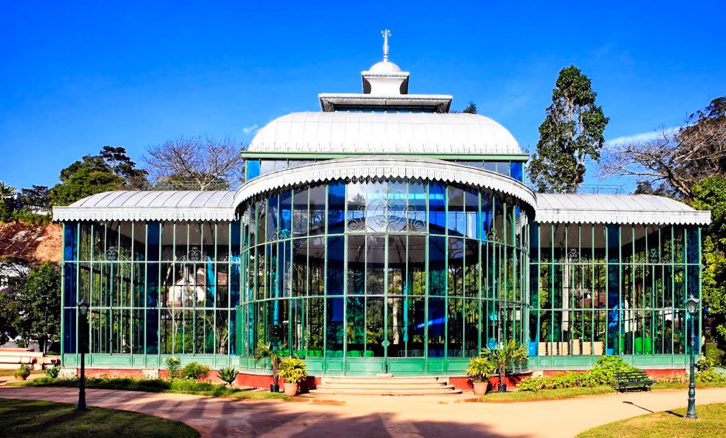A historic glass and iron greenhouse with a domed roof, inspired by Colonial Highlights, stands under a clear blue sky. Its arched windows reflect greenery reminiscent of the Brazil Coffee Region and trees in the background.