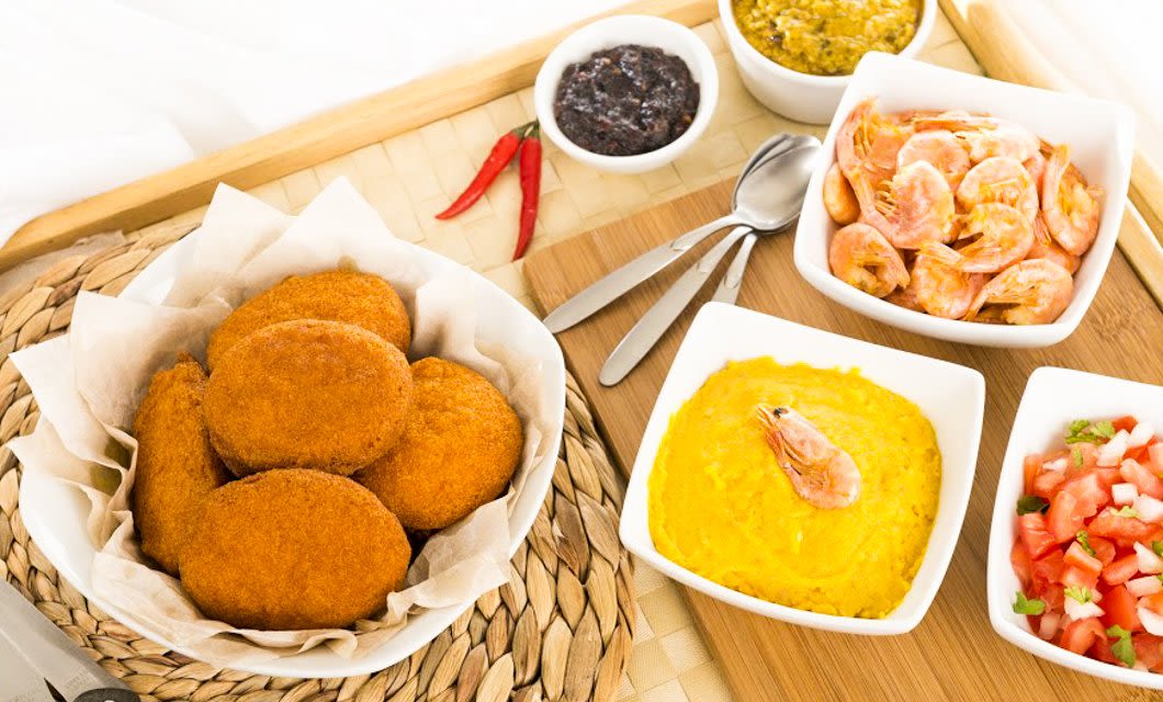 A table with a basket of fried patties, bowls of yellow dip with shrimp, shrimp in sauce, diced tomatoes, and condiments on a wooden tray highlights delicious Brazilian cuisine, with utensils and red chili peppers nearby.