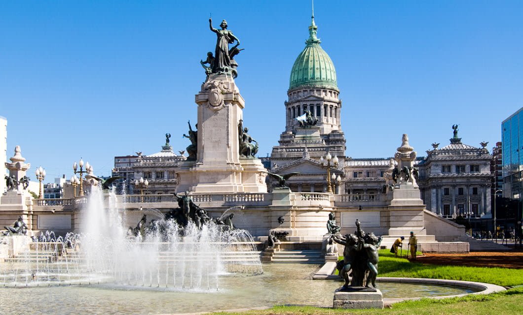 A large neoclassical building with a green dome stands behind an ornate fountain and statues in a public square, evoking the grandeur of Argentina under a clear blue sky.