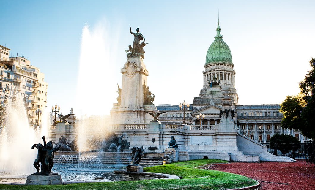 A grand historic building with a green dome stands behind ornate statues and fountains, water spraying into the air, surrounded by a landscaped park in bright daylight—a striking scene in the land of contrast: Argentina.