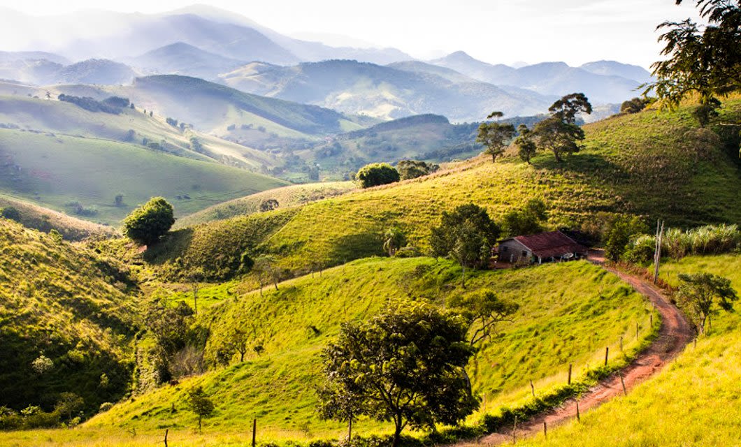 A winding dirt road leads to a small house nestled among green hills and trees, with rolling mountains beyond—just footsteps from the Brazil Gold Route, where a historic train once traced paths beneath the bright sky.