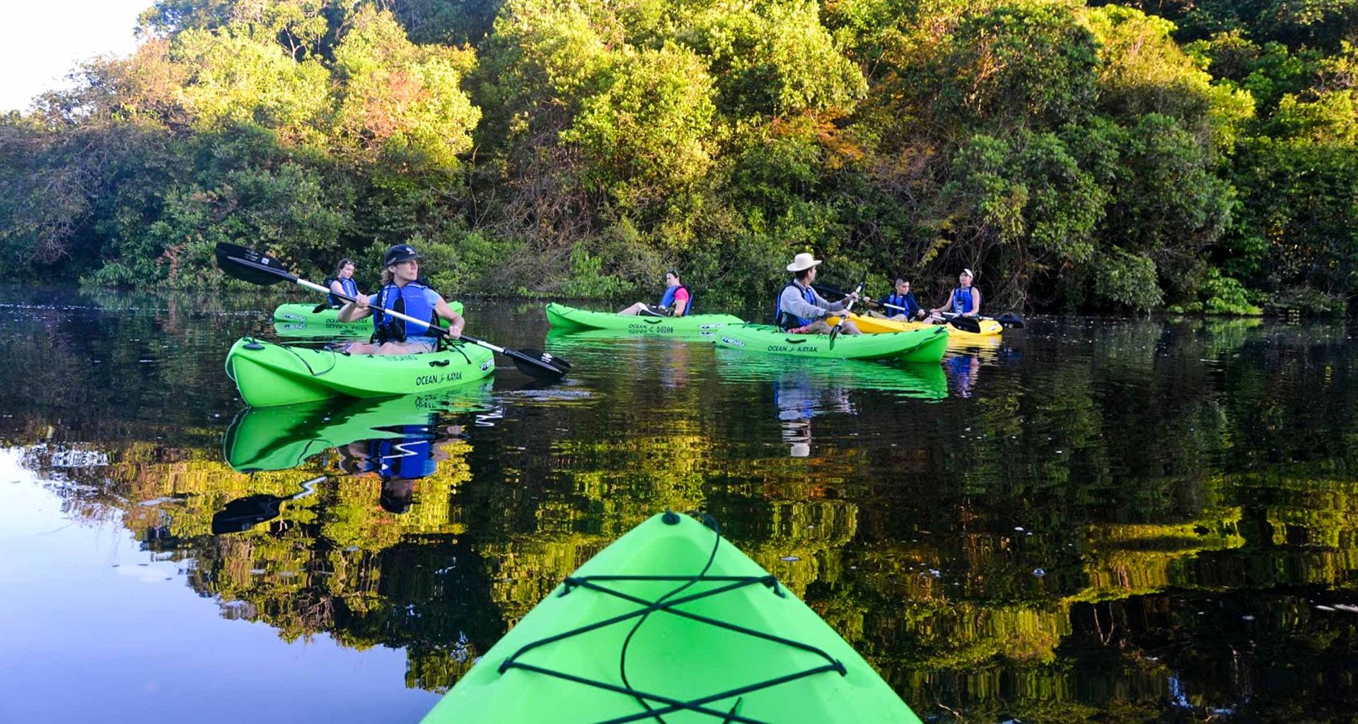 A group of people kayaking on calm water surrounded by lush green trees in the Atlantic Rainforest, with kayaks and foliage reflected on the surface. The photo is taken from the perspective of someone in a green kayak.