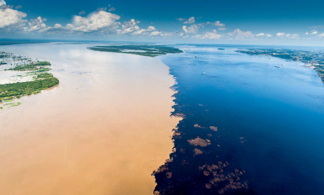 Aerial view of the Meeting of Waters in Brazil Amazon, where the muddy brown water of the Amazon River meets the dark blue Rio Negro, creating a sharp dividing line under a bright blue sky.