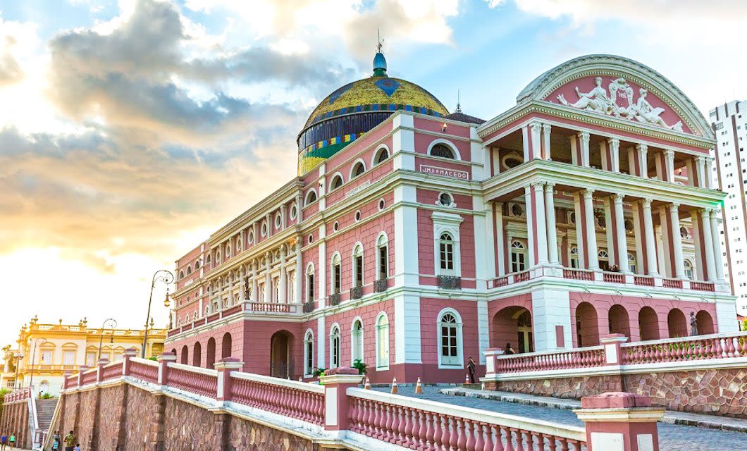 Pink and white neoclassical building with ornate columns and a large, colorful dome, photographed at sunset during a South America Tour. The structure sits elevated with stone steps and railings; sunlight streams through partly cloudy skies.