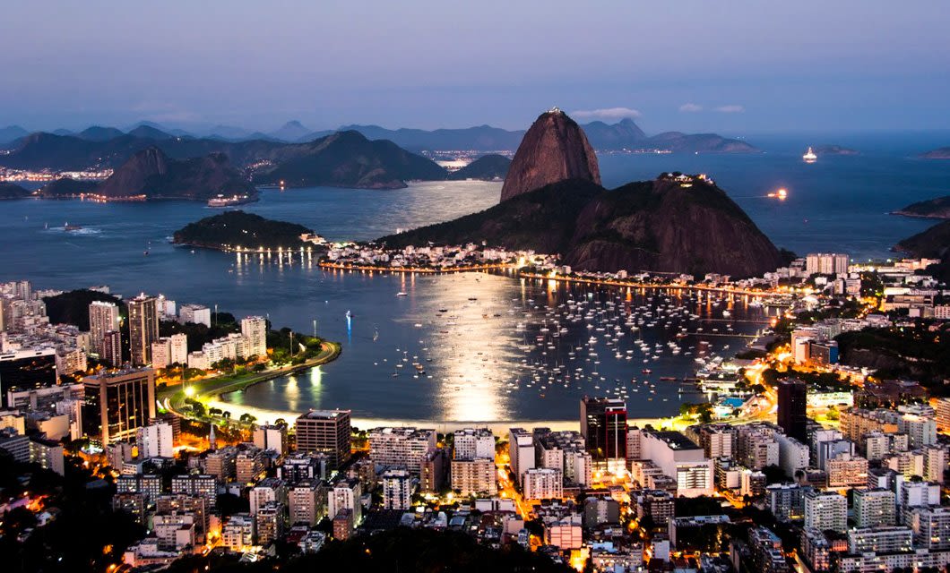 A panoramic view of Rio de Janeiro at dusk, with city lights illuminating buildings and boats in the bay—an unforgettable sight often included on a Rio de Janeiro tour, with Sugarloaf Mountain rising amid hills and water.
