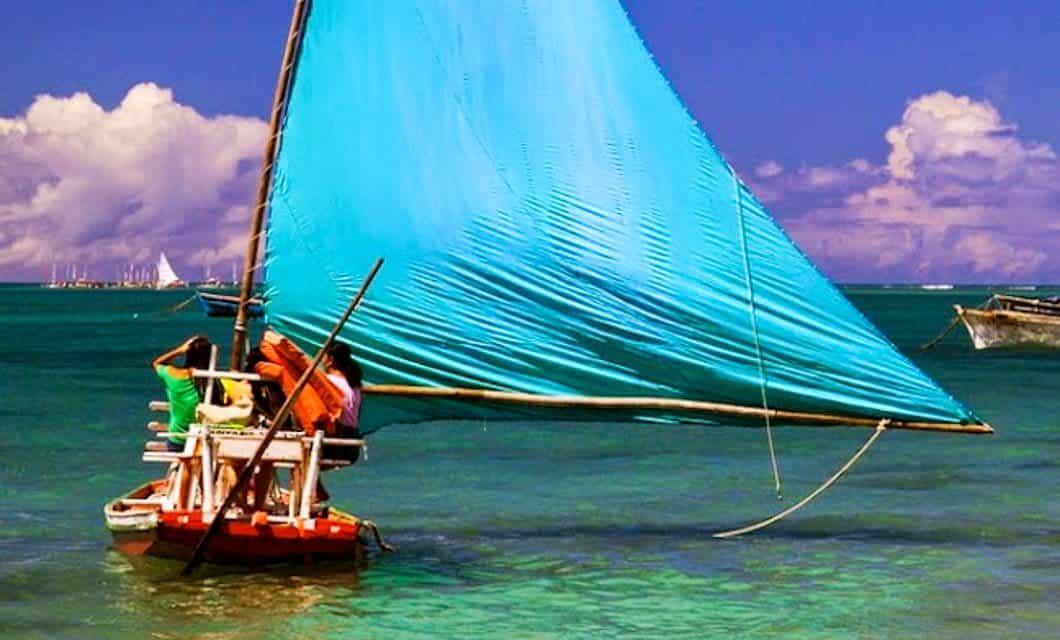 A small wooden sailboat with a large bright blue sail glides on clear turquoise water under a sunny sky along the Coast of the Conquerors. Two people are on the boat, and distant sailboats are visible on the horizon.