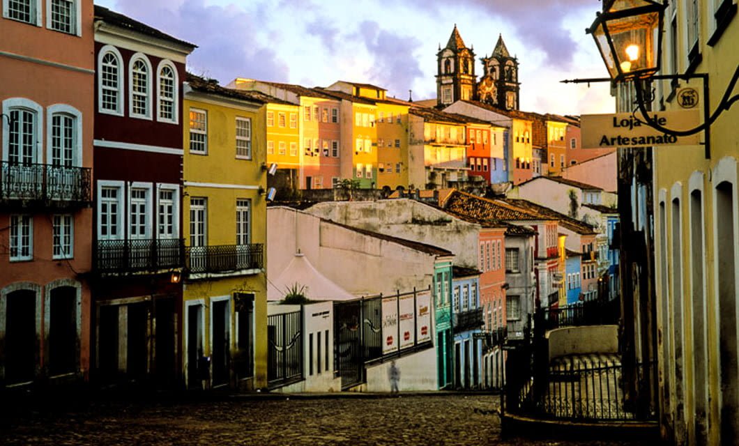 A cobblestone street lined with colorful colonial buildings leads uphill toward a historic church at sunset in Pelourinho, Salvador, Brazil, capturing the charm of the Coast of the Conquerors. A street lamp and craft shop sign are visible on the right.