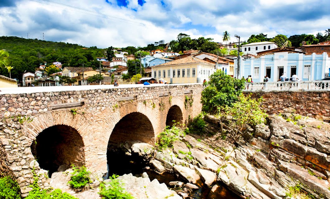 A historic stone arch bridge crosses a rocky riverbed in front of colorful colonial-style buildings and lush green hills, inviting hikers to explore the scenic trails of Chapada Diamantina under a partly cloudy sky.