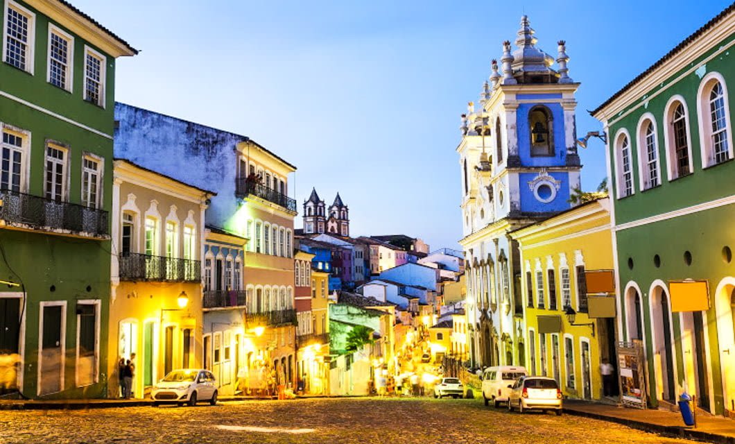 A vibrant street scene in Pelourinho, Salvador, Brazil, captures the charm of a South America Tour with colorful colonial buildings, ornate churches, cobblestone roads, and people strolling along the sidewalk at dusk.