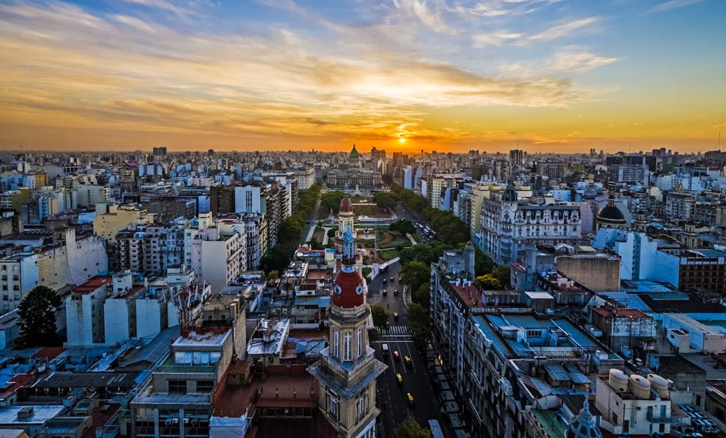Aerial view of a cityscape at sunset, with the sun setting behind distant buildings, a wide avenue lined with trees, and the historic Tucuman-Buenos Aires Railway weaving through a mix of old and modern architecture under a vibrant sky.