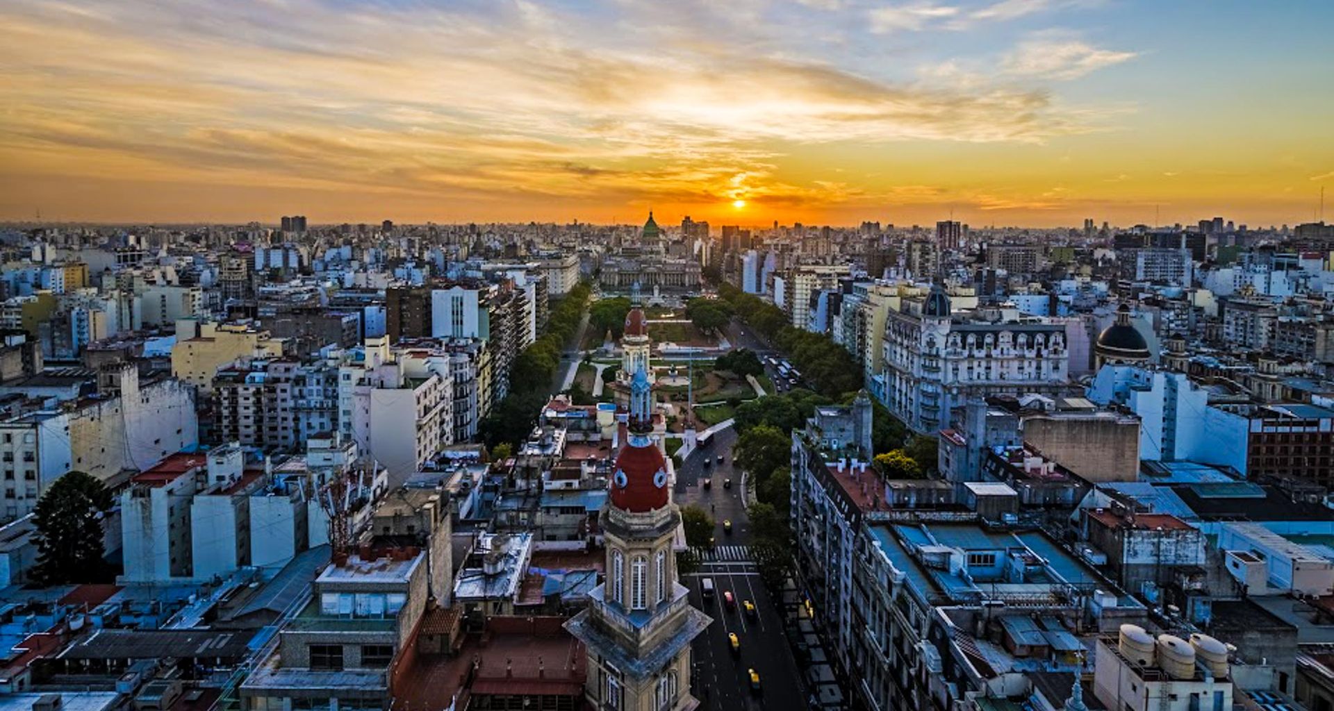 Aerial view of a city with a park-lined avenue leading to a domed landmark at sunset, surrounded by densely packed buildings under a colorful sky—a scene reminiscent of the Tucuman-Buenos Aires Railway journey.
