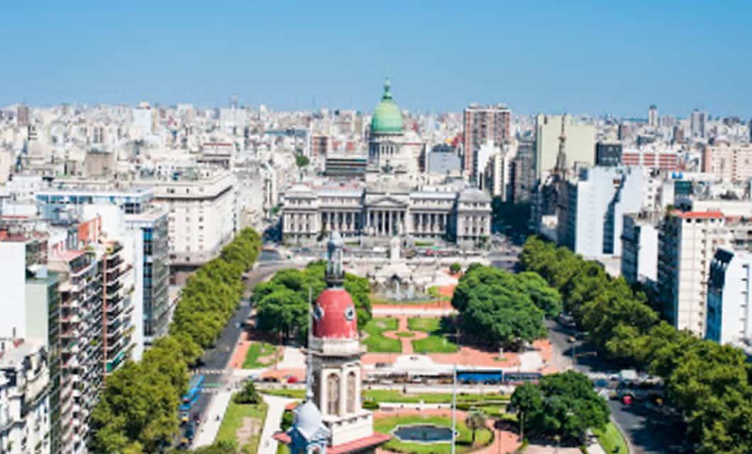Aerial view of Buenos Aires showcasing a green park, wide boulevards, and a prominent domed government building in the center, surrounded by tall buildings under a clear blue sky.