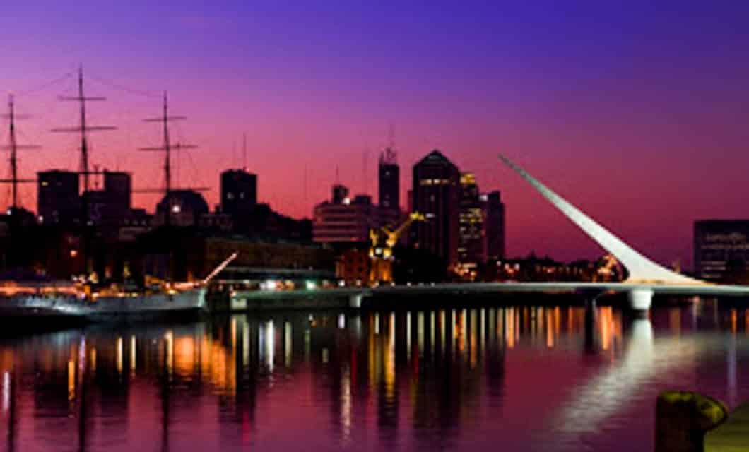 Cityscape at dusk with a modern white bridge spanning a calm river in Argentina, tall ships docked along the waterfront, and colorful lights reflecting in the water. High-rise buildings are silhouetted against a purple and pink sky.