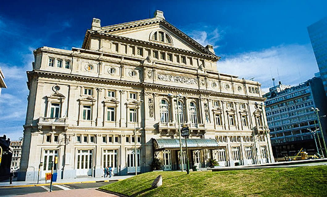 A grand, historic opera house with ornate architectural details, arched windows, and columns stands under a clear blue sky in Argentina. People walk near the entrance, and a manicured lawn is in the foreground.