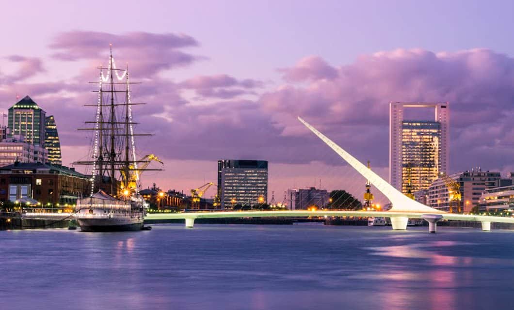 A modern cityscape at dusk features a white, asymmetrical bridge over a calm river, a tall sailing ship docked nearby, and high-rise buildings under a purple sky—evoking the spirit of a grand railway journey like the Tucuman-Buenos Aires Railway.