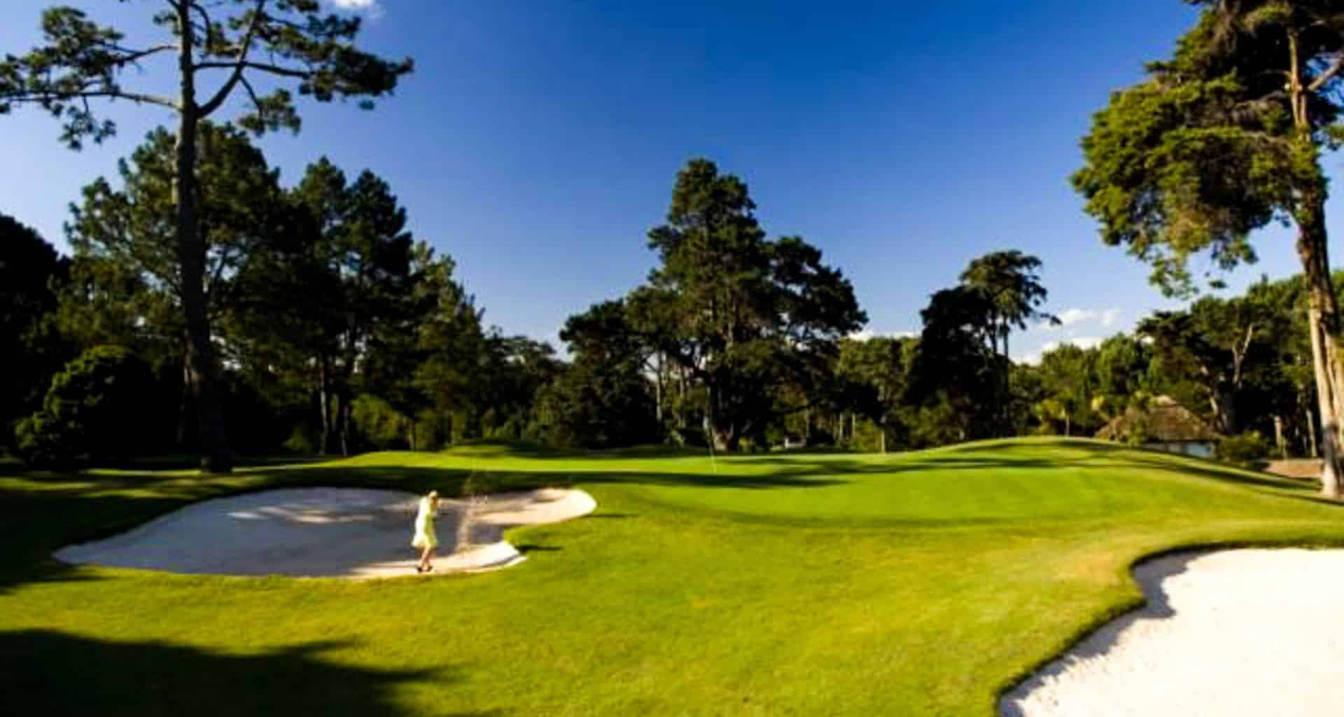 A person stands in a sand bunker on a sunny Argentina Golf course, surrounded by lush green fairways, large trees, and a clear blue sky.
