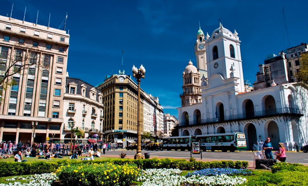 A busy South American city square with a white colonial-style building, flowerbeds, people walking, and several buses on the street, set against a clear blue sky and surrounding historic buildings—perfect for starting a wine tour adventure.