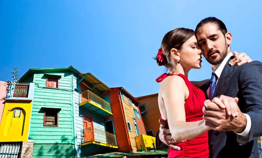 A couple dances tango in front of colorful buildings under a clear blue sky in Buenos Aires. The woman wears a red dress with a red flower in her hair, and the man wears a dark suit.