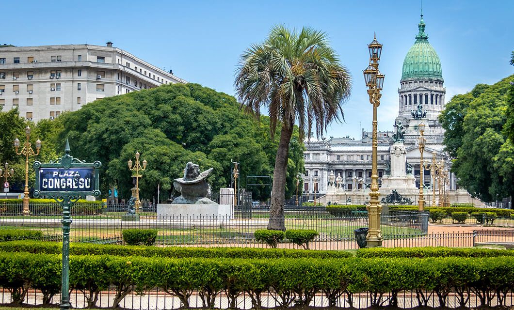 A landscaped plaza with trimmed hedges, a palm tree, ornate street lamps, sculptures, and a sign reading Plaza Congreso. The Coast to Coast Tour lets you admire the grand building with its striking green dome in the background.
