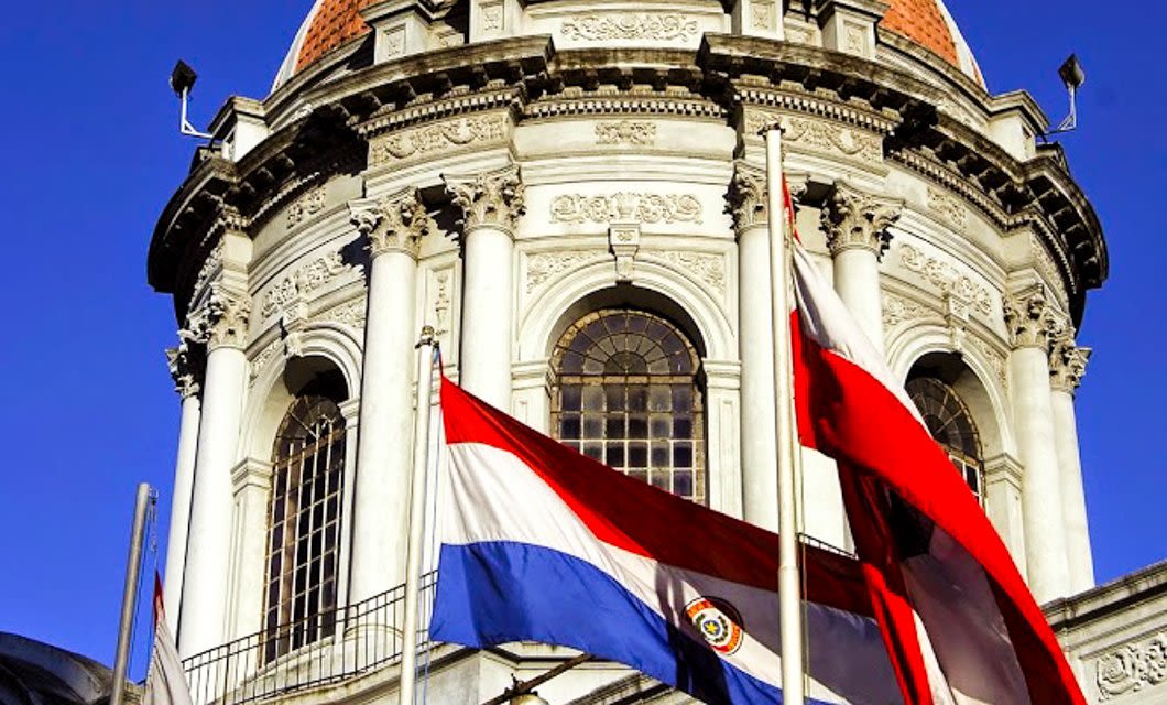 A close-up of a neoclassical building with tall arched windows and columns, topped with a dome. In the foreground, the flags of Paraguay and another country wave—an iconic sight during any Paraguay Tour. The sky is clear and blue.