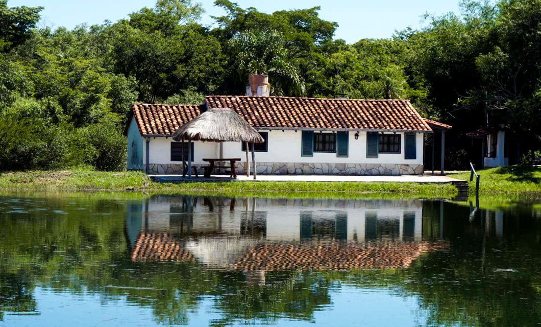 A small white house with a red-tiled roof stands by a calm pond in the lush greenery of Chaco. Its reflection shimmers in the water, and a thatched gazebo with a picnic table sits nearby.