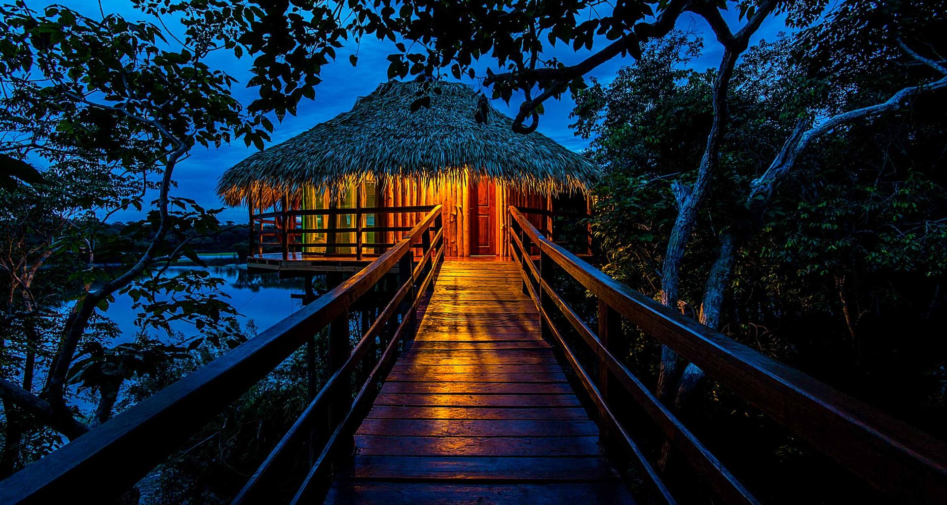 A thatched-roof hut on stilts is warmly lit at night at Juma Amazon Lodge, seen from a wooden walkway surrounded by dense Amazon rainforest and reflected in the water below.