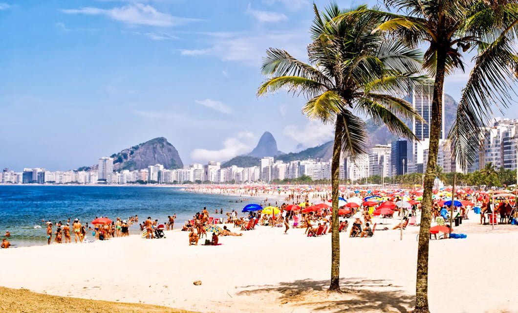 A crowded beach in Rio de Janeiro with people sunbathing and relaxing under colorful umbrellas, palm trees in the foreground, and city buildings with mountains in the background on a sunny day—a perfect spot for a Beaches Tour.