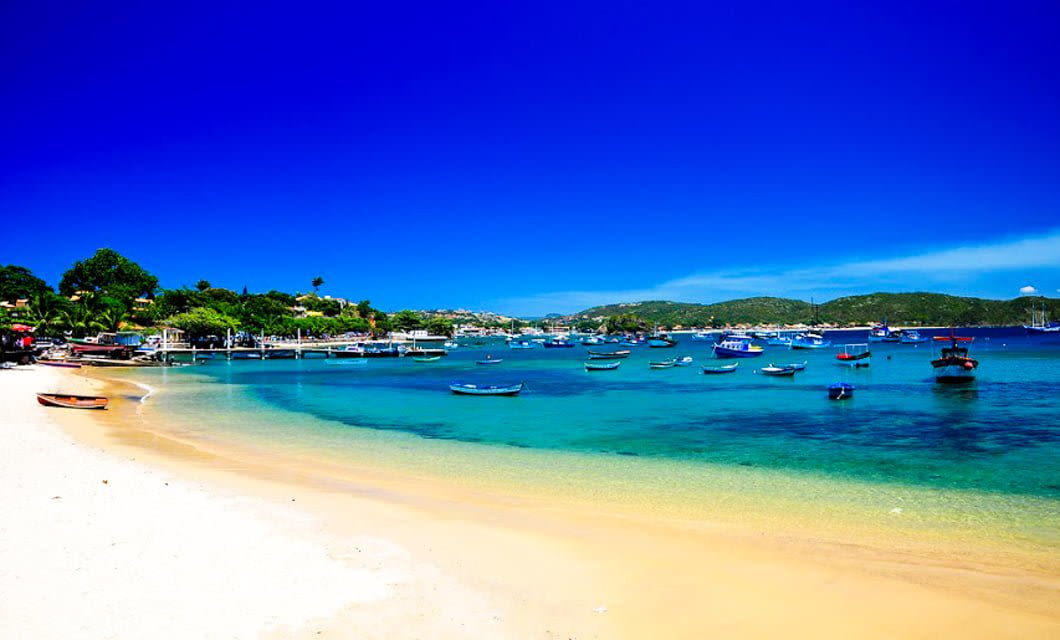 A bright, sunny beach scene on Buzios Beaches near Rio de Janeiro, with clear turquoise water, white sand, and small boats anchored by the shore. Green hills and buildings rise in the background under a deep blue sky—perfect for a memorable tour.