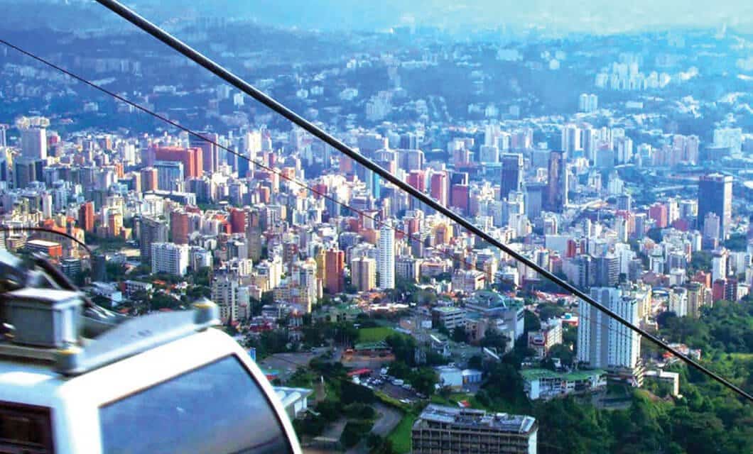A cityscape with numerous high-rise buildings surrounded by hills and greenery, viewed from a cable car ride—one of the Venezuela highlights—cables and part of the cabin visible in the foreground.