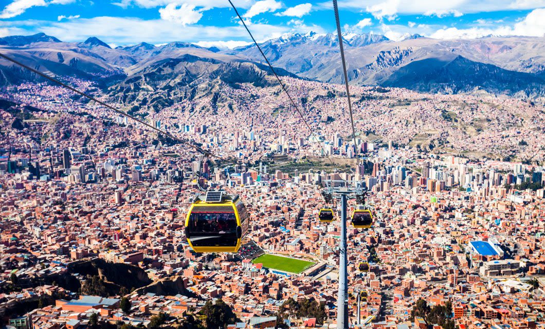 Aerial view of colorful cable cars on a Coast to Coast Tour gliding over the densely packed city of La Paz, Bolivia, with mountains in the background under a partly cloudy sky.