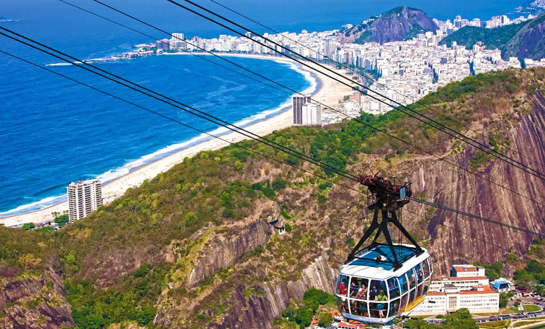 A cable car glides above green hills toward Salvador Bahia, where white buildings line a curved coastline and sandy beach, with the blue ocean and distant mountains completing this stunning Brazil tour scene.