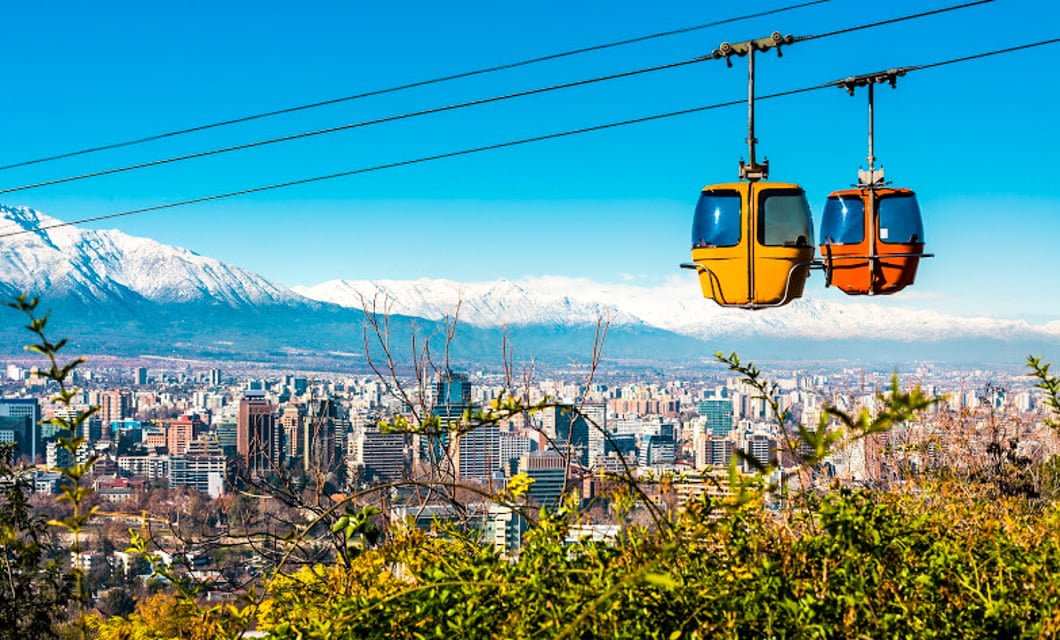 Two colorful cable cars travel over a city with tall buildings, greenery, and snow-capped mountains in the background under a clear blue sky—an inspiring view for wine experts seeking new tasting destinations.