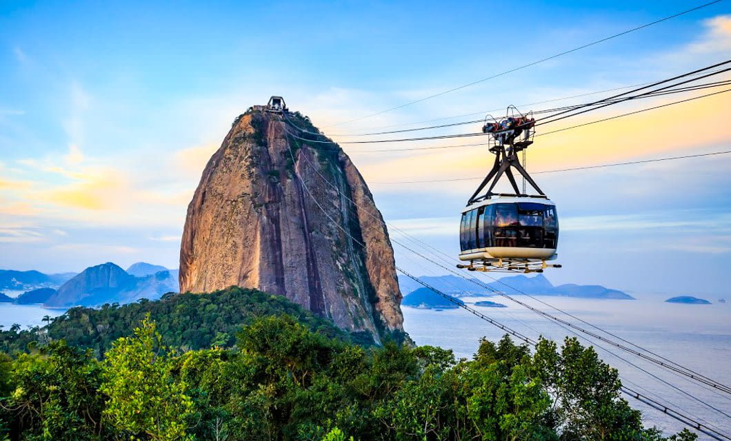 A cable car travels toward Sugarloaf Mountain, part of the Brazil Gold Route, rising above green trees and overlooking the ocean in Rio de Janeiro under a colorful sunset sky.