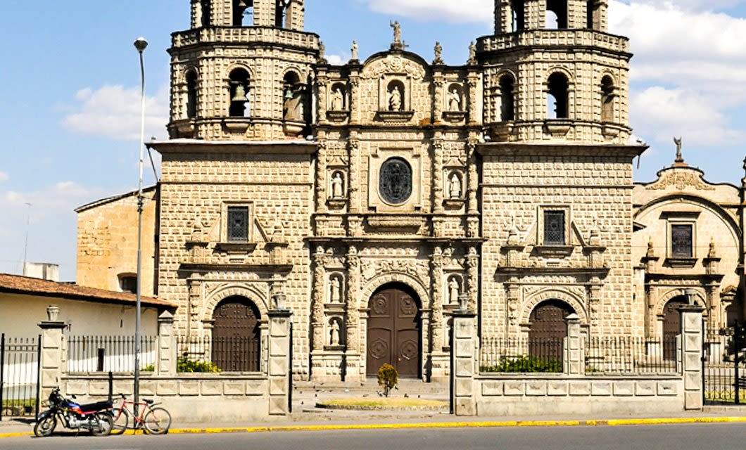 A historic stone church in Cajamarca with two bell towers, elaborate carvings, large wooden doors, and a small garden in front; a motorcycle is parked near the sidewalk outside the fence.